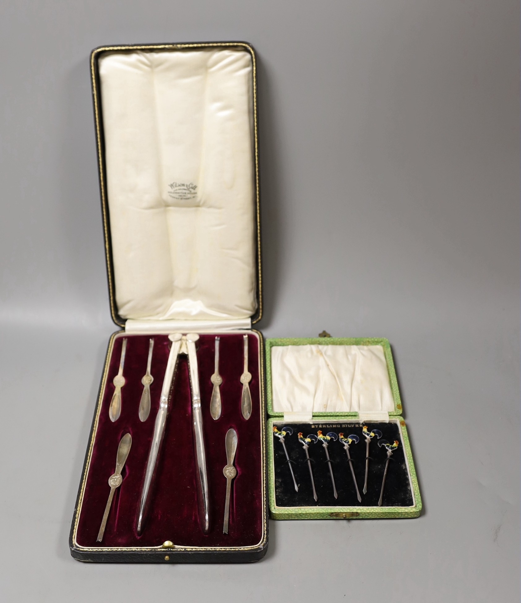 A George V cased set of six silver lobster picks and plated crackers and a similar set of silver and enamel cocktail sticks